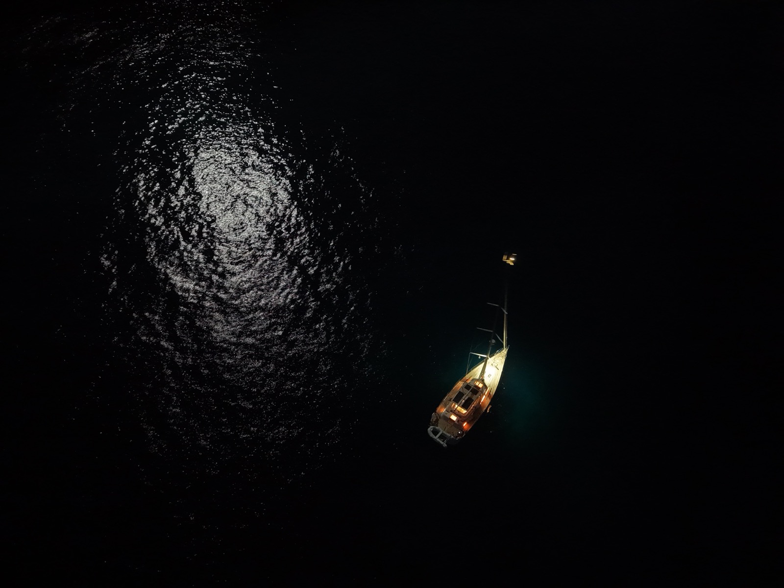 Drone shot from above of Stani lit up at anchor at night off Sardinia's southwest corner, moonlight on the water beside her